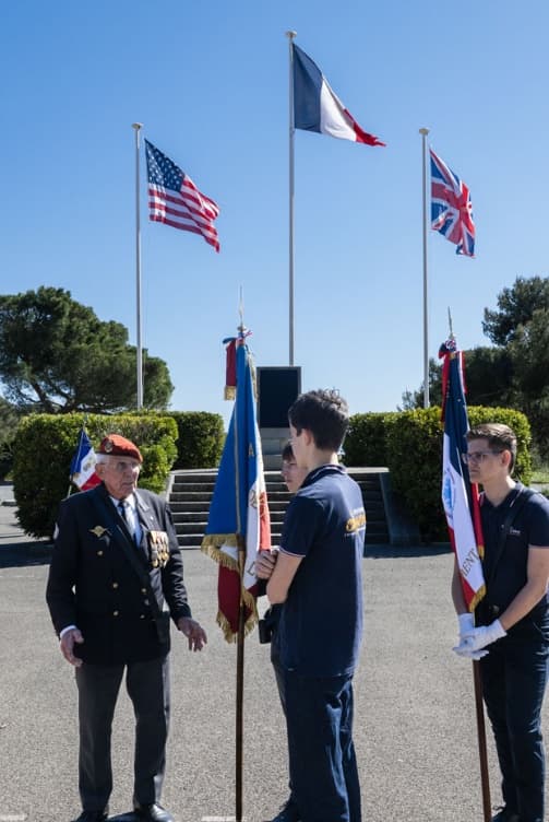 Un ancien combattant échange avec de jeunes porte-drapeaux devant les drapeaux alliés